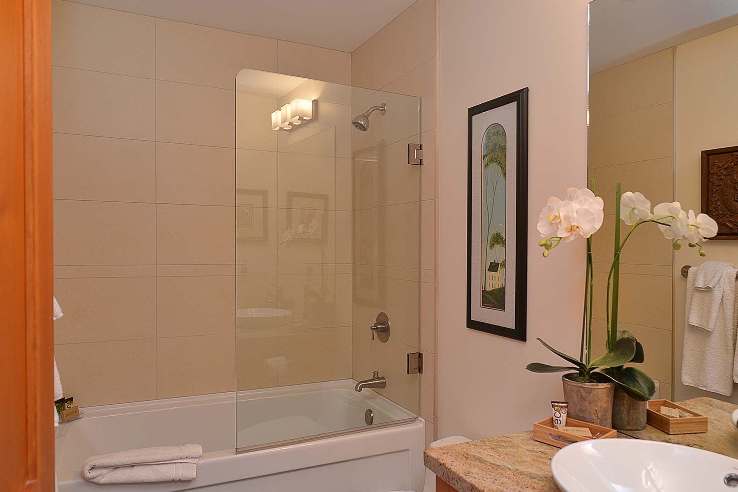 One of the bathrooms of this vacation house rental has a shower with a connected bathtub, artwork on the wall, and flowers.